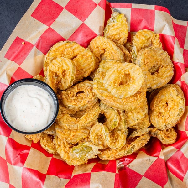A basket of golden-brown fried pickles with a side of creamy ranch dressing on red and white checkered paper.