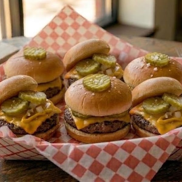 Six cheeseburger sliders with pickles and diced onions in a red and white checkered paper-lined basket.